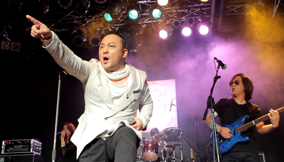 Singer on stage points outward during a live concert, with guitarist and drums in the background.