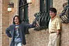 Two men stand by a brick building with iron window grilles and wall lanterns.