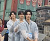Three young performers in clear ponchos pose outside a traditional building with decorative banners.