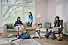 Several young adults sit and lie around a living room with a sofa, large windows and casual items on the floor.