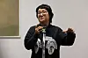 Artist speaking into a microphone during a briefing at a museum, with a framed artwork visible behind.