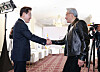 Two men shake hands in a bright room as photographers capture the moment.