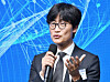 Lee Hae-jin speaks into a microphone at an AI summit with a blue digital background.