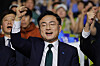 Political rally scene with a man in a suit raising both fists in front of a crowd at an indoor convention.