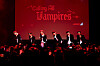 Performers on a red-lit stage in front of a crowd and a backdrop that says 'Calling All Vampires.'
