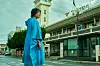 Woman in a blue robe standing on a street near a historic building with columns and a tower.