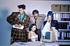 Four people gathered around a vintage desktop computer in an office with binders and paperwork.