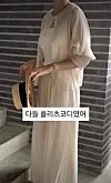 Woman in a cream pleated dress and cardigan stands by a brick wall holding a straw hat.
