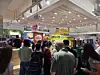 People crowd around a brightly lit booth inside a terminal with colorful promotional signs.