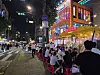 Crowd dining outdoors next to a brightly lit building on a city street at night.