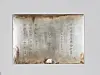 Faded rectangular board with vertical writing and rust marks mounted on a gray wall.