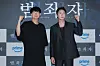 Two men in dark outfits pose with raised fists in front of a step-and-repeat backdrop.