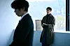 Takigawa stands indoors near a large window with a blurred man in the foreground.