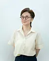 Woman with short hair and glasses wearing a light shirt stands against a plain background.