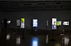 Dark gallery room with several illuminated vertical screens standing on black pedestals.
