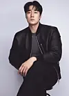 Man seated against a light background wearing a black jacket and dark outfit.