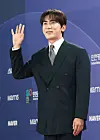 Kim Won-hoon waves in a dark suit on a red carpet against a blue event backdrop.
