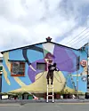 Person painting a large abstract mural on the side of a blue building while standing on a ladder.