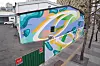 Abstract blue, green and white mural painted on a building next to a tree-lined walkway.