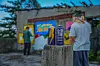 Two people painting a mural outdoors with spray paint cans on a concrete post in front.