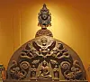 Intricately carved circular reliquary with figures, topped by a sculpted head, against a golden wall.