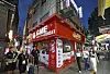 A red GME Electronics storefront on a busy nighttime street with pedestrians walking nearby.
