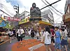 Crowds move through a busy market street beneath a giant gorilla statue and overhead wires.