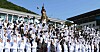 Large group of Navy cadets in white uniforms cheer and throw caps into the air outdoors.