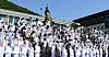 Large group of Navy cadets in white uniforms cheer and throw caps into the air outdoors.