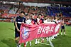 Football players stand on the field holding a red banner and greeting the crowd in a stadium.