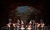 Ballet dancers perform on a stage with a red forest backdrop, statues, and a central staircase.