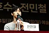 Jeon Min-chul speaks into a microphone at a press conference with a nameplate and water bottle in front of him.