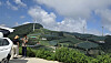 People taking photos beside a car with green terraced hills and wind turbines under a blue sky.