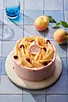 A pink frosted cake with fruit topping on a round board, next to peaches and a blue glass on tiles.
