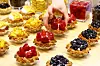 Hands arrange strawberry tarts on a table lined with colorful fruit pastries at a bakery fair.