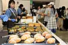 A bakery booth with trays of assorted bread and pastries as visitors look on at a trade fair.