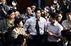 Man in tie answers questions while surrounded by reporters and cameras in a crowded media gathering.