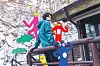 Lee Chae-min in a teal jacket on a wooden outdoor structure with colorful illustrated figures and trees behind him.