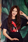 Smiling woman with long dark hair poses in a black graphic T-shirt against a leafy backdrop.