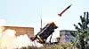 A Patriot missile launches diagonally upward from a ground-based launcher amid smoke and trees.