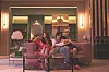 Two people sit on a sofa in a warmly lit, upscale lounge with flowers and a glass coffee table.