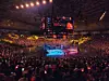 Packed arena surrounding a brightly lit central stage during a live event.