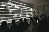 Visitors watch a bright cascading-line installation in a dark exhibition space.