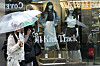 Shop window displaying fall knitwear and mannequins as pedestrians with umbrellas pass outside.