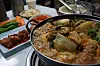 Bubbling skillet of seafood and vegetables beside colorful side dishes on a restaurant table.
