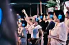 A crowd in a dark venue wears blue-lit headphones, with several people raising their hands.