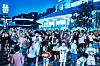 Large nighttime crowd raising hands and phones outside a brightly lit building.