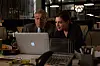 Two colleagues work behind an Apple laptop at a desk with a phone and office supplies.
