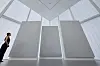 A visitor stands beside three large textured gray artworks in a bright modern museum gallery.