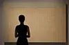 A silhouetted museum staff member stands before a large textured beige artwork.